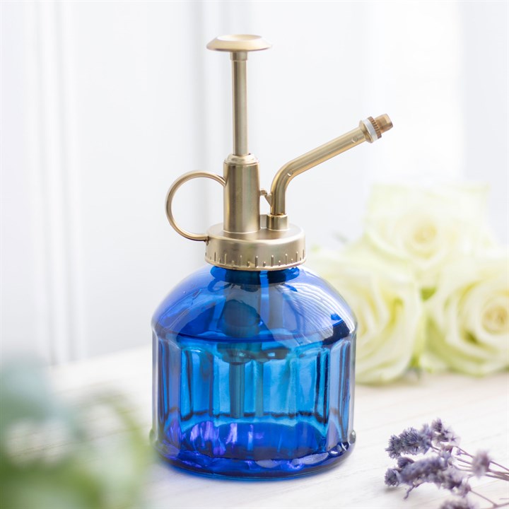 Blue glass spray bottle with brass nozzle on a white surface with flowers in the background