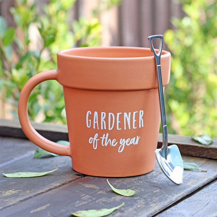 Terracotta mug with 'Gardener of the Year' text and garden trowel on a wooden surface with greenery in the background