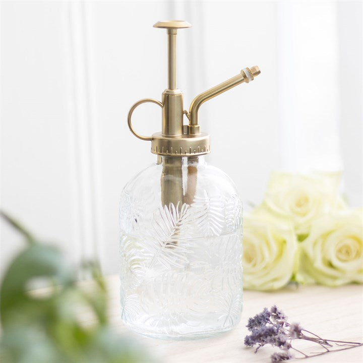 Clear glass spray bottle with brass nozzle on a light background with flowers and plants.