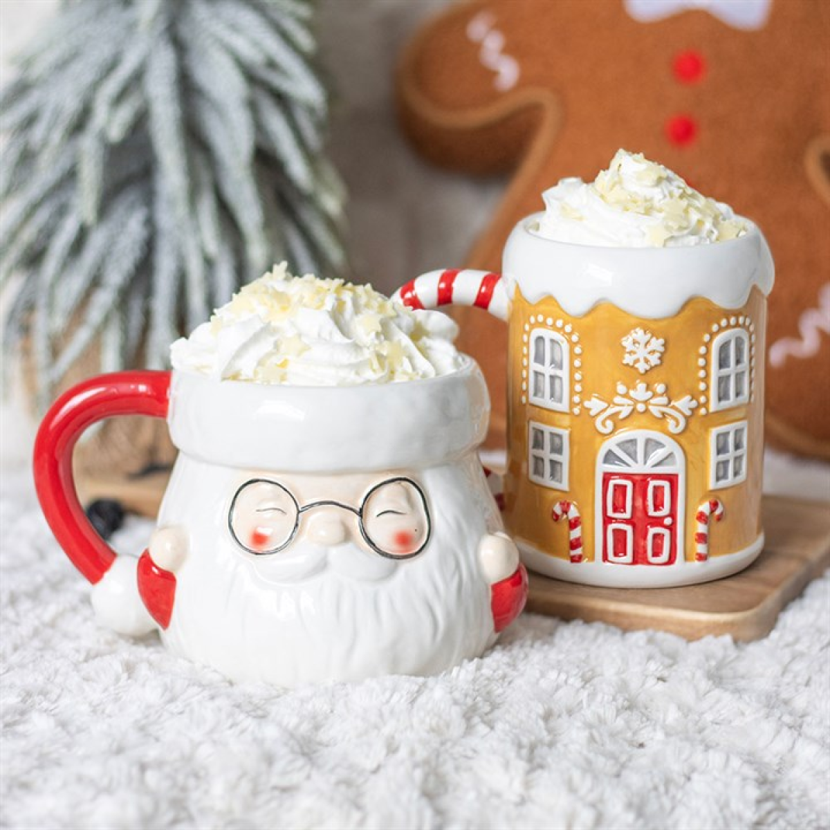 Two mugs with whipped cream; one shaped like a Santa face, the other like a gingerbread house, on a snowy surface with a gingerbread man in the background.