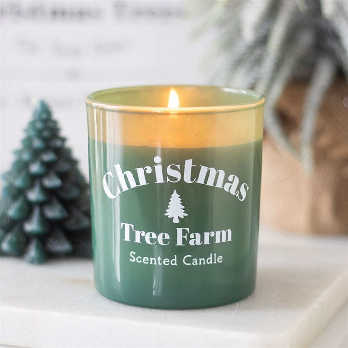 Green candle labeled 'Christmas Tree Farm Scented Candle' with a gold lid, placed on a surface with a blurred background.