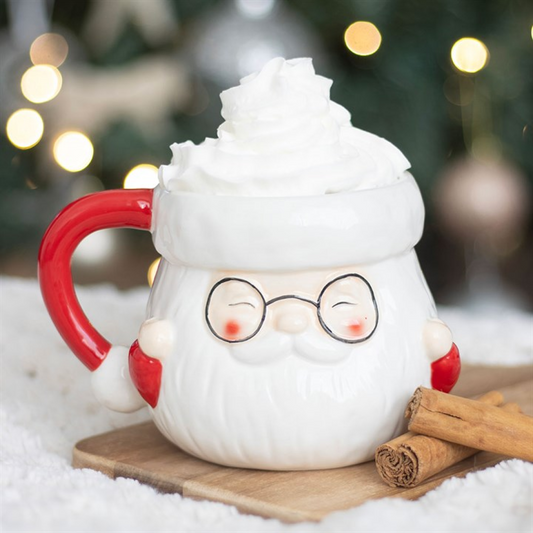 White mug with a cartoon face, red handle, and glasses, set against a festive background with lights and cinnamon sticks.