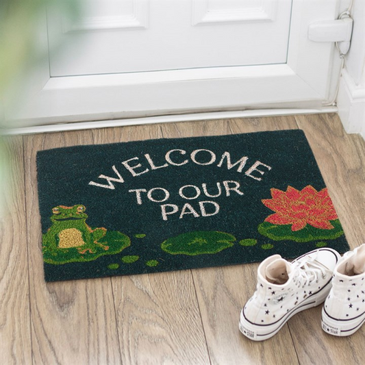 Doormat with frog and lily pad design and 'Welcome to our pad' text on a wooden floor.