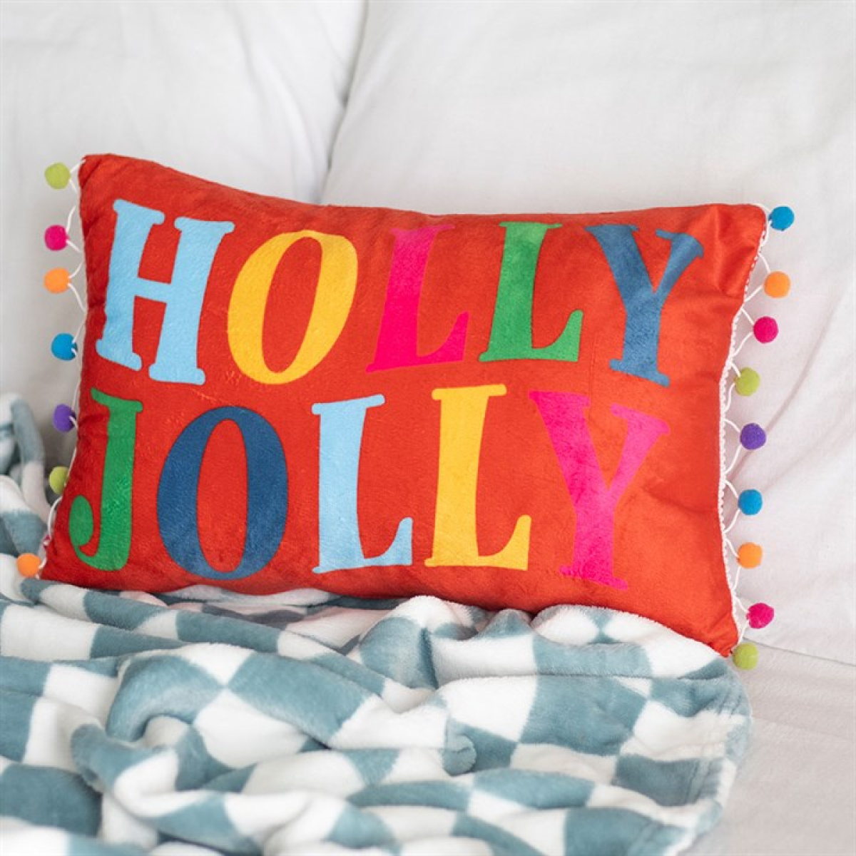 Colorful 'Holly Jolly' pillow on a patterned blanket with pom-poms.