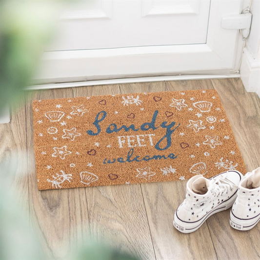 Doormat with 'Sandy Feet Welcome' text on a wooden floor with white sneakers.