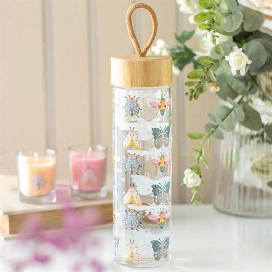 Glass water bottle with bamboo lid and animal illustrations on a surface with candles and flowers in the background.