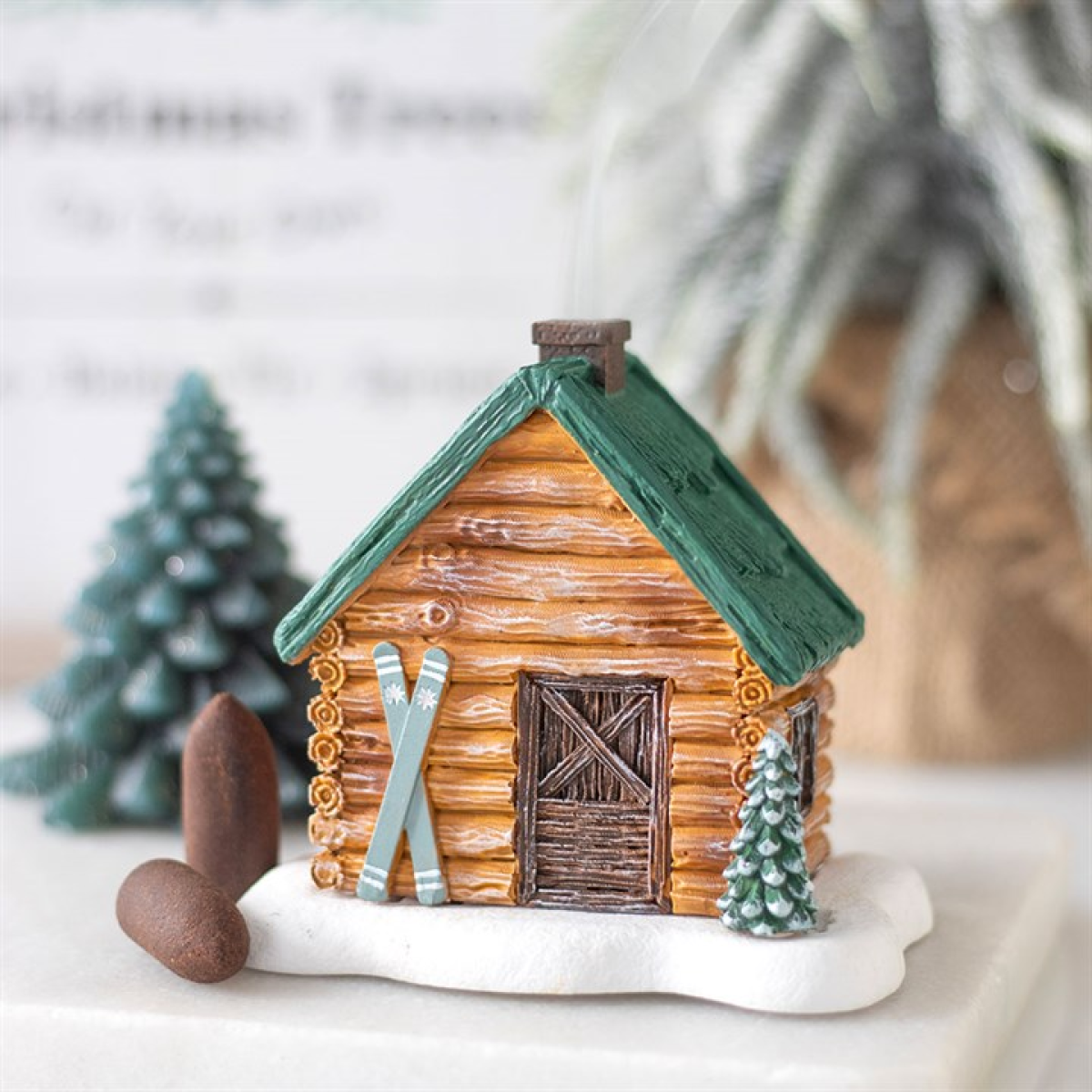 Small log cabin figurine with green roof and skis on a white surface with decorative trees.