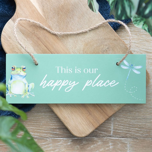 Decorative sign with a frog illustration and text 'This is our happy place' on a wooden surface.