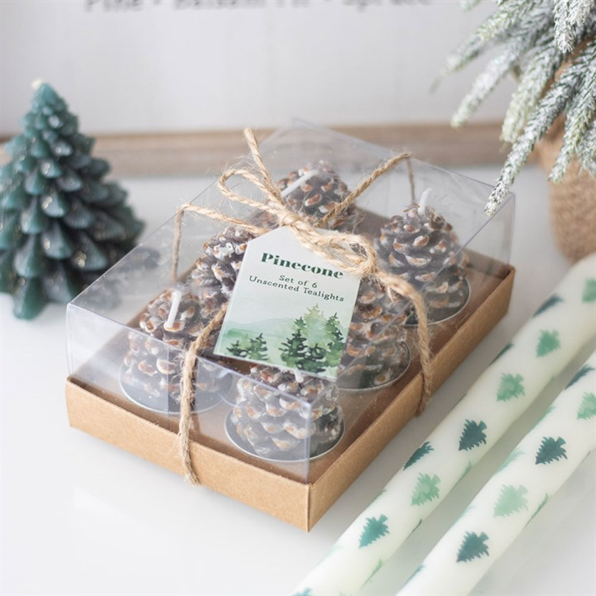 Set of pinecone candles in a clear box with a decorative tag, surrounded by Christmas-themed wrapping paper and small trees.