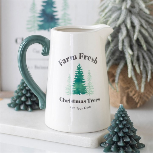 White ceramic pitcher with 'Farm Fresh Christmas Trees' text and tree graphics on a surface with decorative trees.